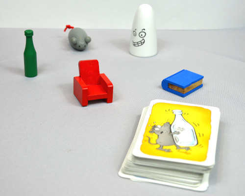 Im Kreis stehen 5 Holzfiguren: ein weißer Geist, ein roter Sessel, ein blaues Buch, eine grüne Flasche und eine grüne Maus. Im Vordergrund liegt ein Kartenstapel darauf zu sehen eine Maus mit einer Flasche.